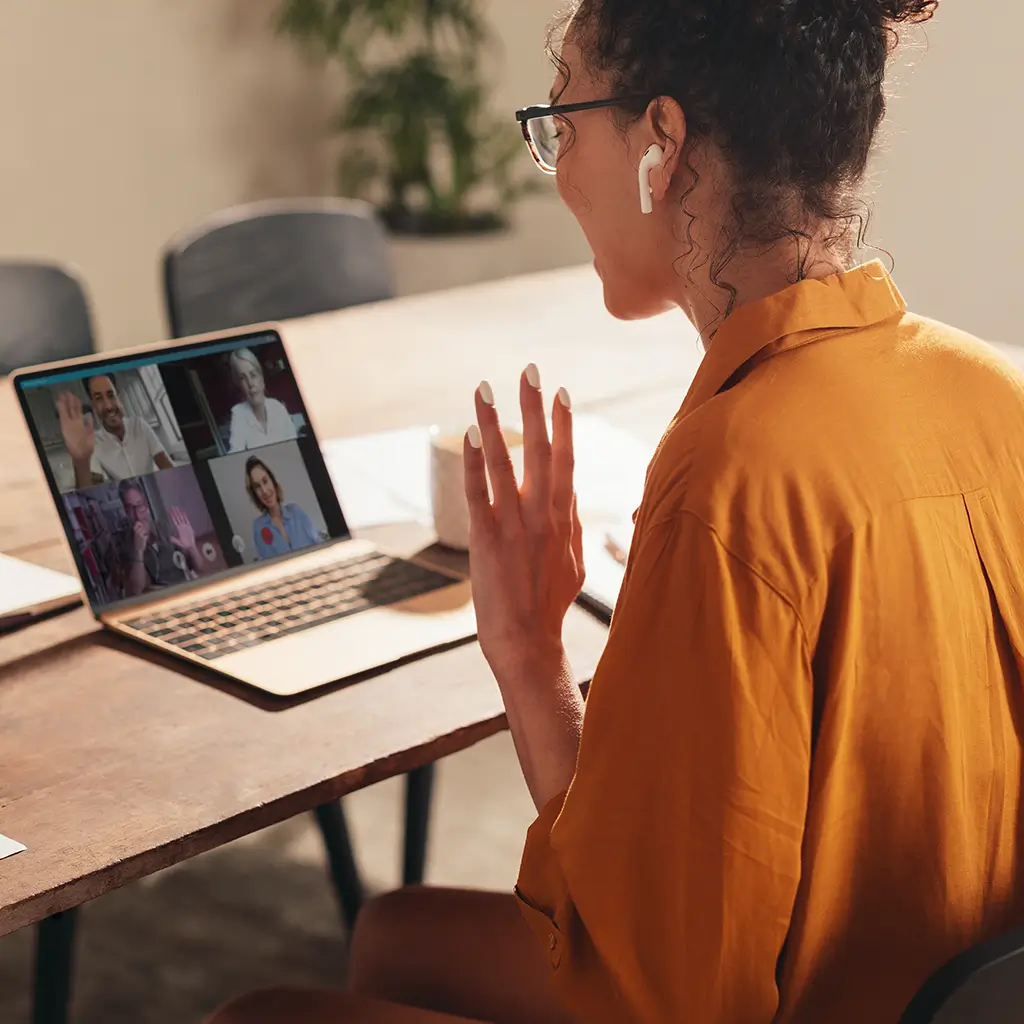 A woman at a table using a laptop, with two people visible on the screen during a video call.